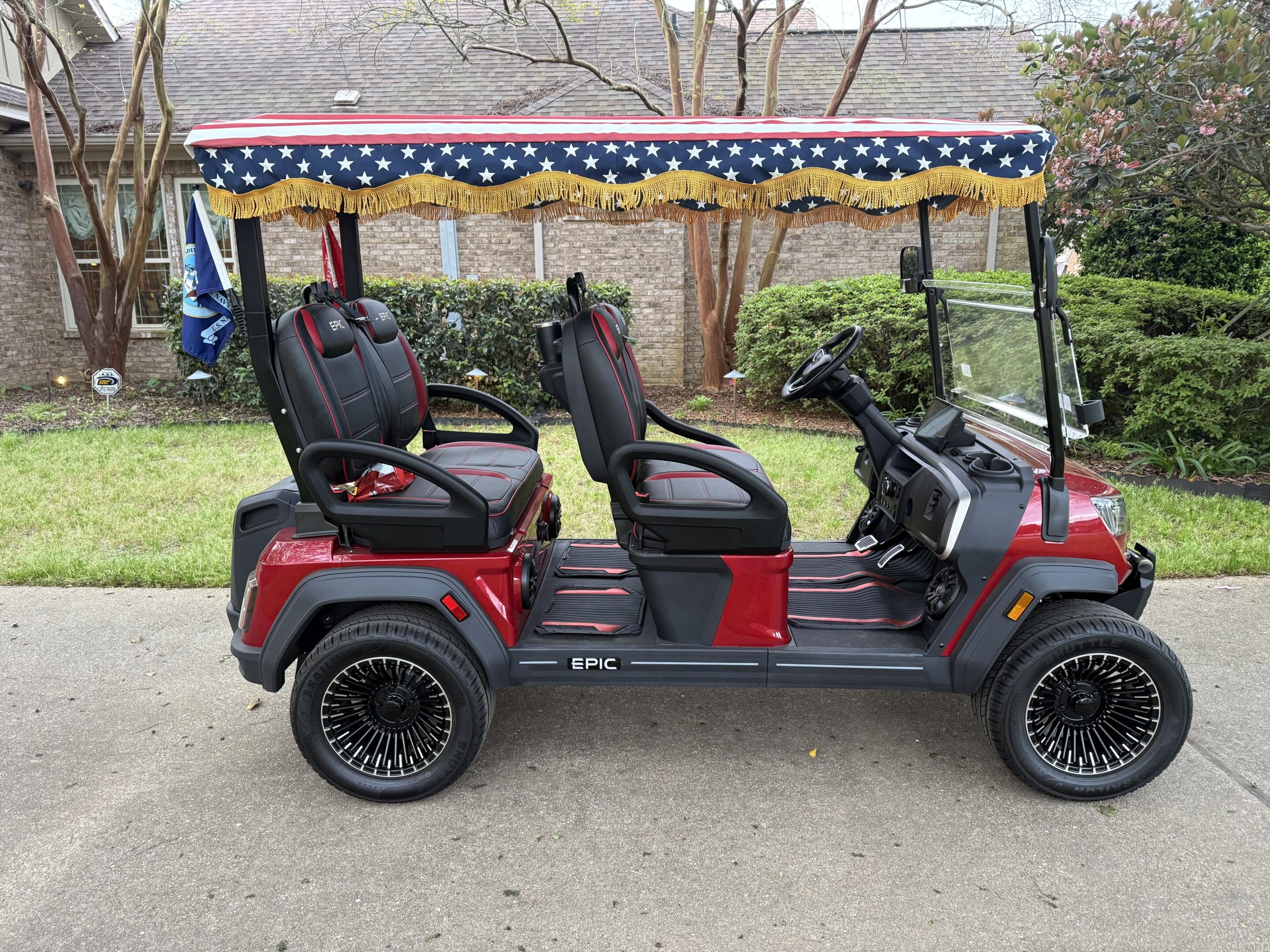 Red golf cart with a star-spangled patriotic canopy and gold fringe, parked on a driveway in front of a brick house.